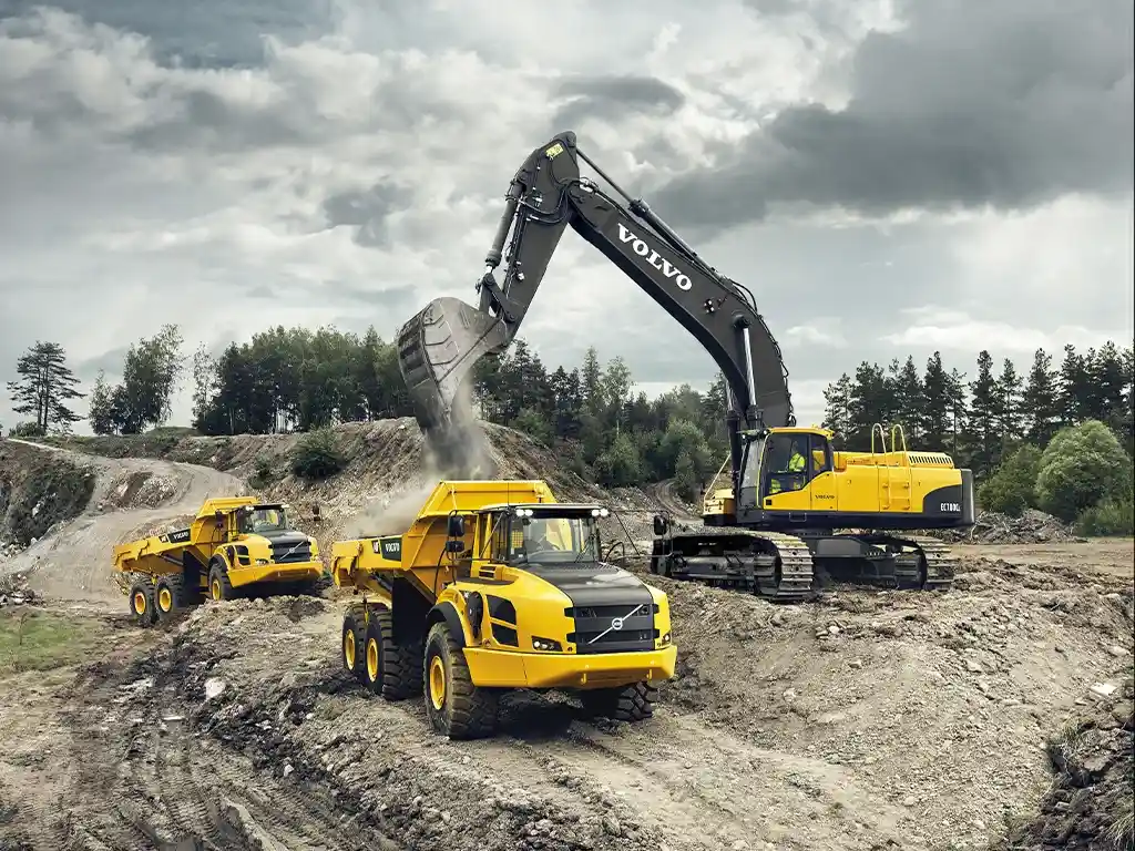 A Volvo excavator loading dirt into two yellow dump trucks at a construction site under cloudy skies