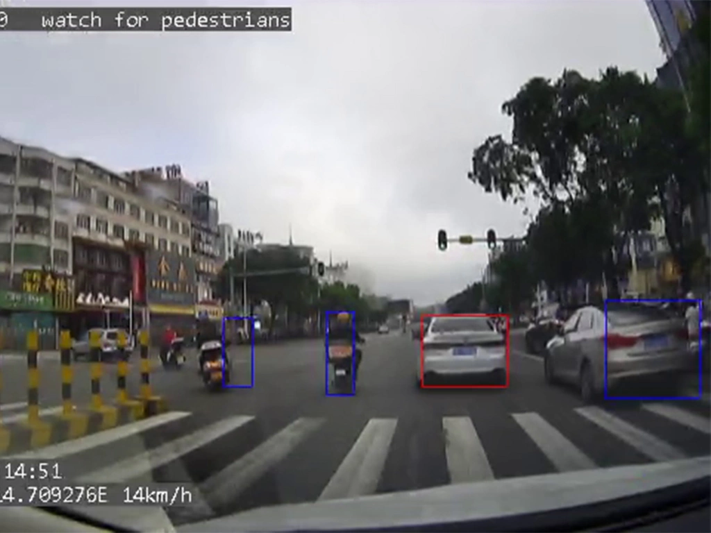 Dashcam view of a city intersection with cars, scooters, and pedestrians in the crosswalk, warning to watch for pedestrians
