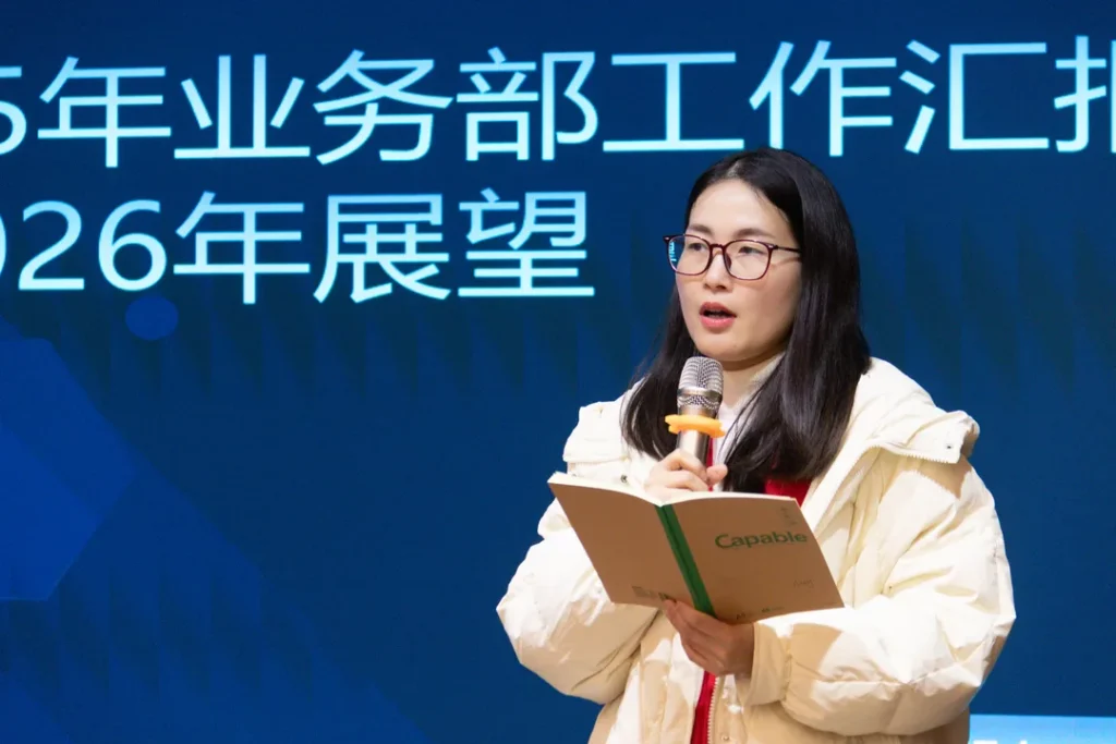 Woman speaking into a microphone while reading from a booklet on stage with Chinese event text behind her