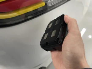 Hand holding a small black electronic module in front of a car bumper