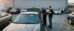 Two men work on wiring inside a parked sedan in a lot surrounded by other cars