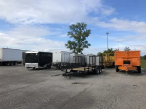 Several utility trailers parked in a lot, including black, white, and orange enclosed trailers under a cloudy sky