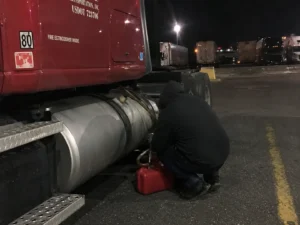 Person crouches beside a semi truck’s fuel tank with a red fuel can at night