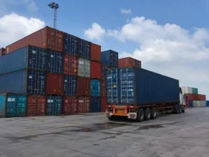 Semi-truck hauling a shipping container beside stacked containers at a port yard