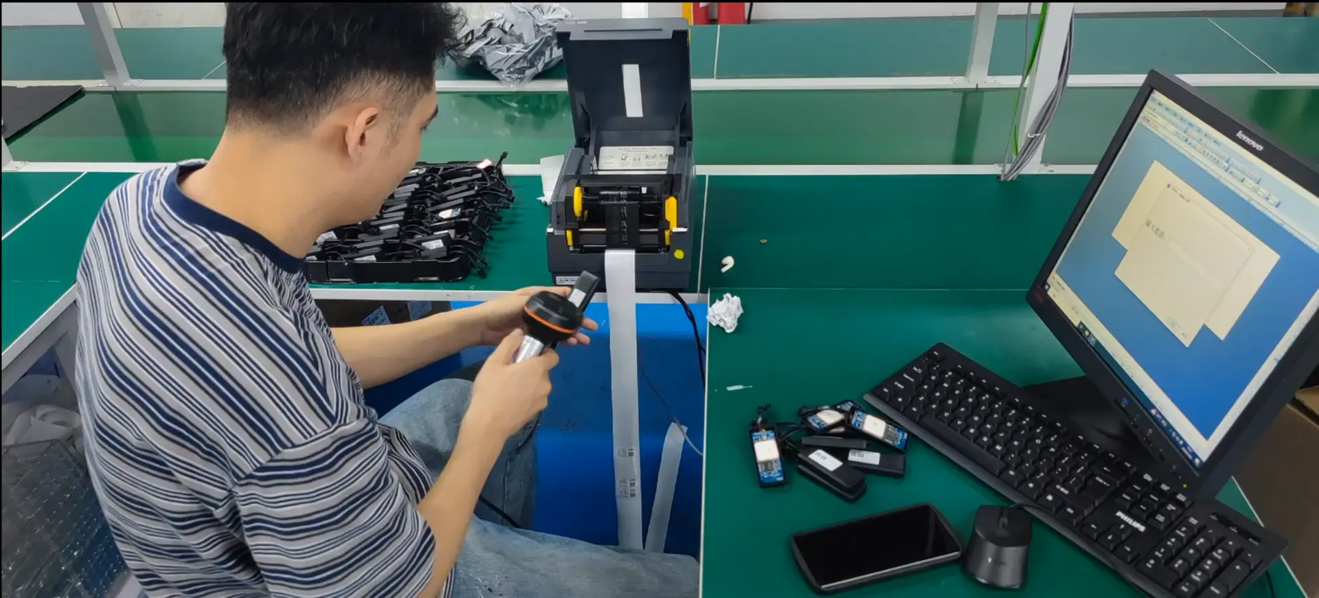 Worker assembling and testing electronic components at a factory workbench with a computer monitor