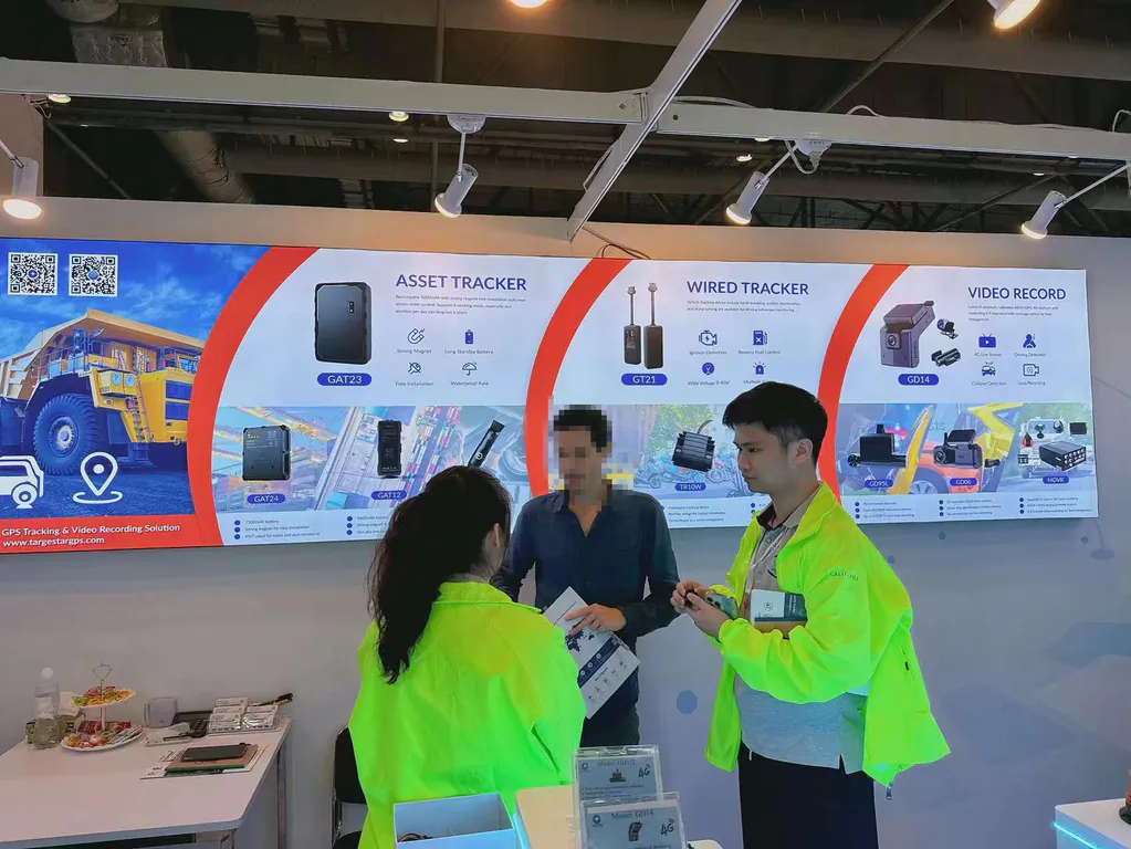 Three people stand at a trade show booth displaying GPS tracking and video recording products